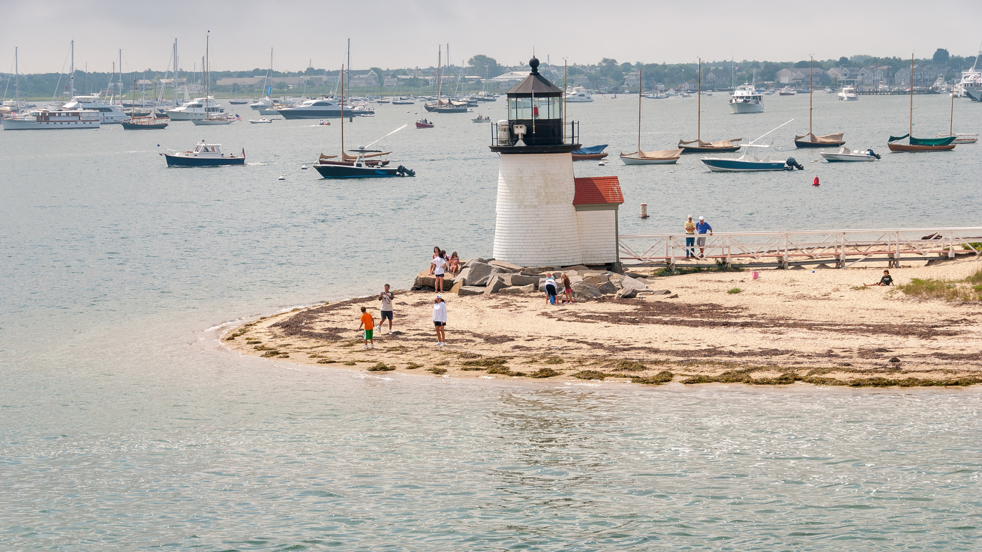 Brant Point Lighthouse Nantucket from the water with sailboats and families on the beach — Sirena Collective