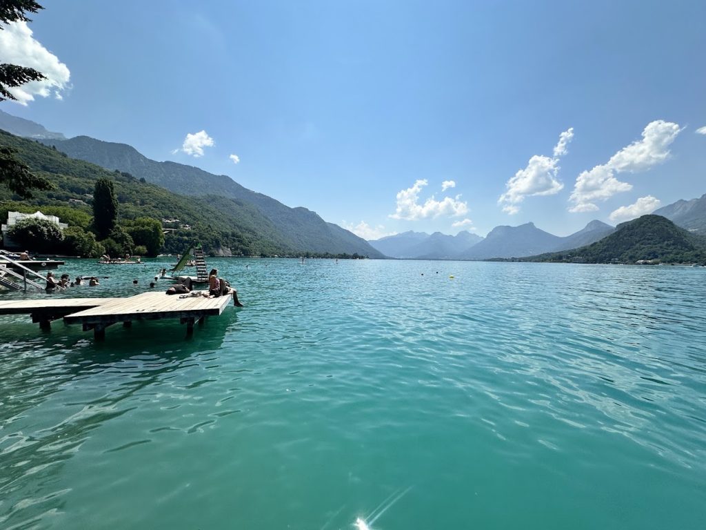 Crystal-clear turquoise waters of Lac d'Annecy with floating dock, swimmers, and the French Alps in the background — Sirena Collective family travel guide